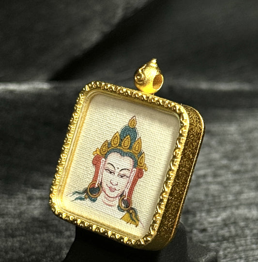 Angled view of a Four-Armed Avalokiteshvara miniature thangka pendant, showing detailed hand-painted deity portrait on textured canvas, natural mineral pigments, and a carved gold-toned square frame with raised ornamental border and hanging loop.