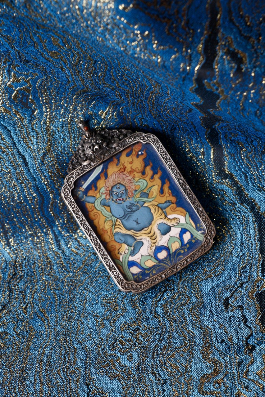 Angled view of a Fudo Myo-o (Acalanatha) thangka pendant on blue-gold brocade, highlighting the wrathful deity in flames and the ornate silver frame.