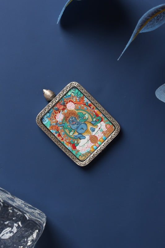 Angled flat-lay of a Fudo Myo-o (Acalanatha) mini thangka pendant in a silver gawu case, showing the fierce blue deity with flaming hair and sword on a dark blue background.