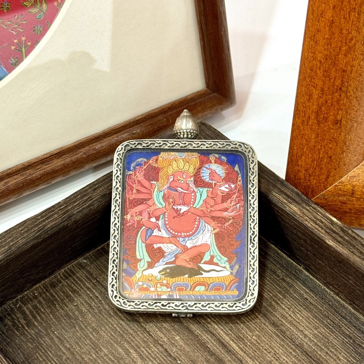 Ganapati (Vinayaka) mini thangka pendant displayed on wooden surface, highlighting the pure silver frame, engraved border pattern, and hand-painted red deity figure.