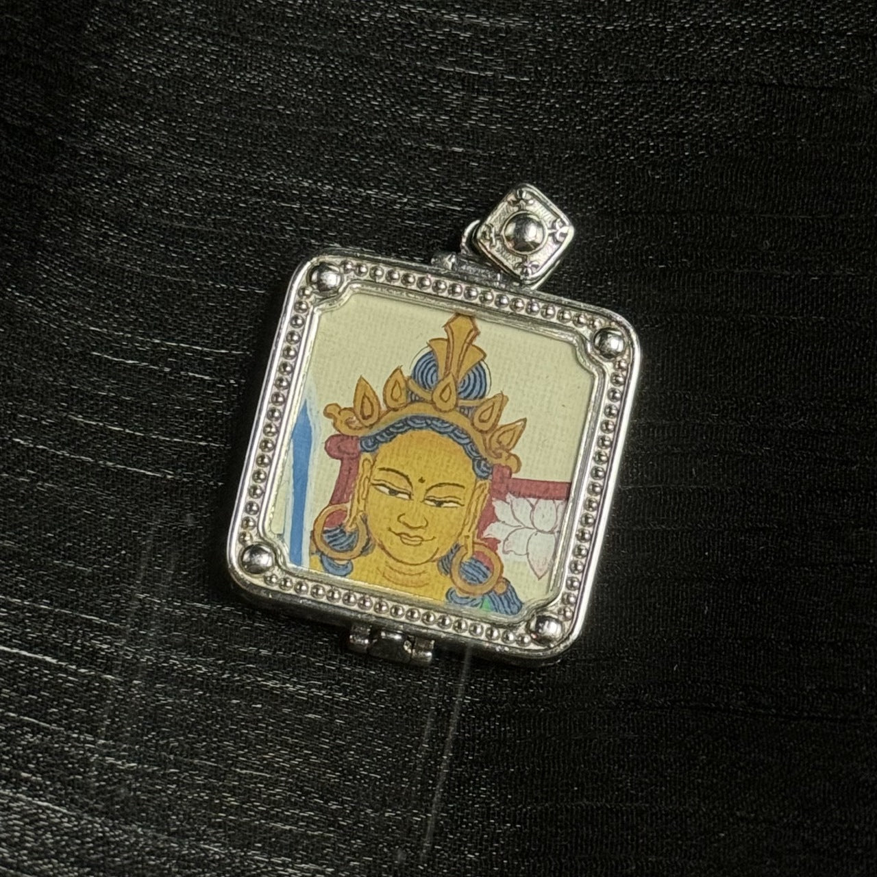 Close-up of a square mini thangka pendant featuring a single-sided hand-painted golden face with a gold crown and blue hair details, with a lotus petal detail visible at the side of the portrait, set in a silver-tone beaded frame with rounded corner beads on black background