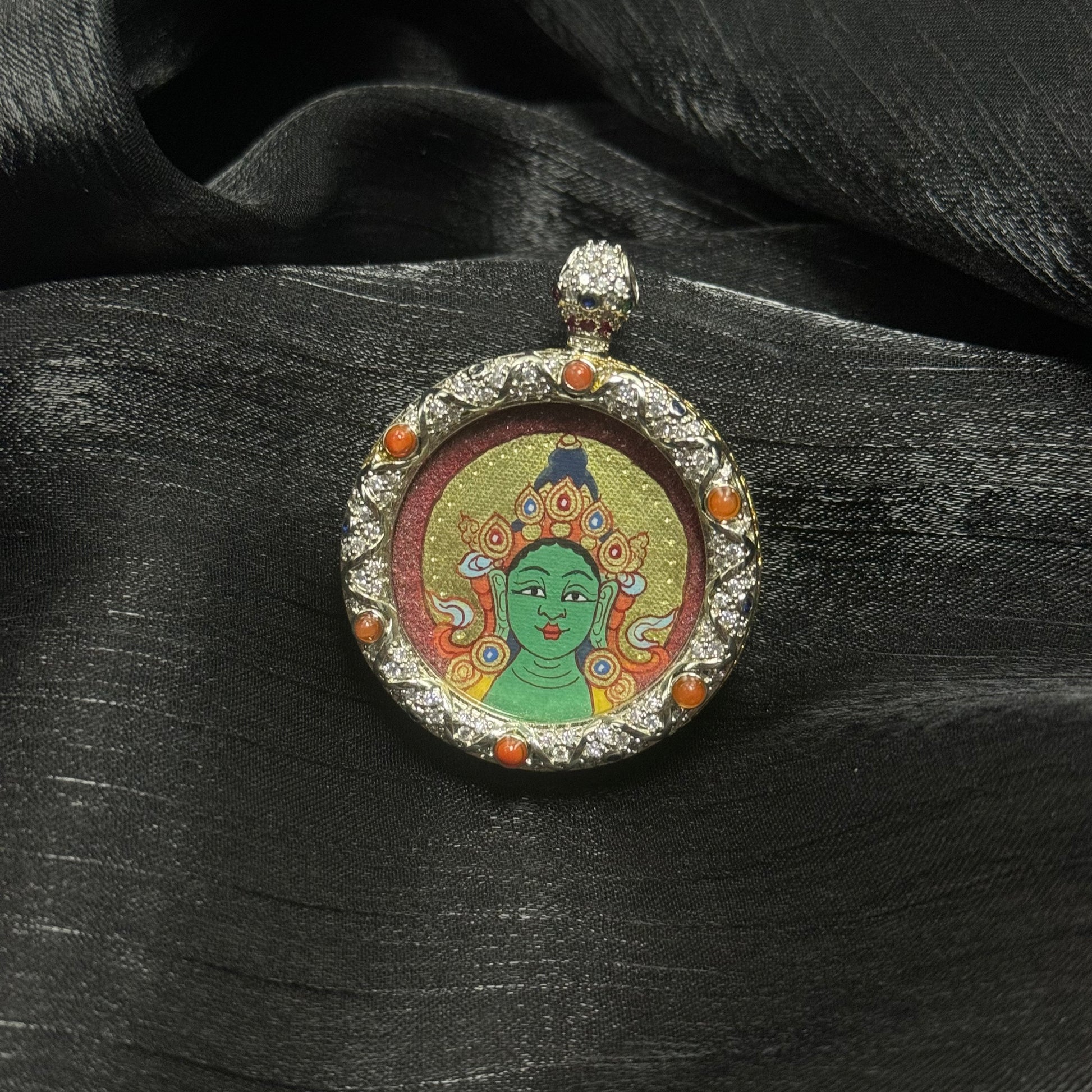 Green Tara miniature thangka painting in traditional Rebgong style with mineral pigments and cinnabar red halo, displayed inside a round jeweled copper pendant case placed on dark fabric background.