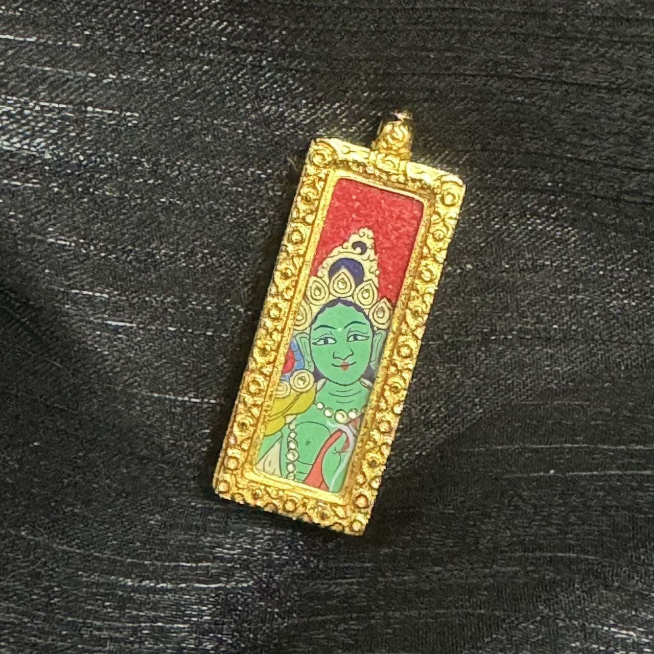 Angled close-up of a Green Tara mini thangka pendant highlighting the raised gold-tone border texture, the red background panel, and the hand-painted facial features and jewelry details within the copper case.