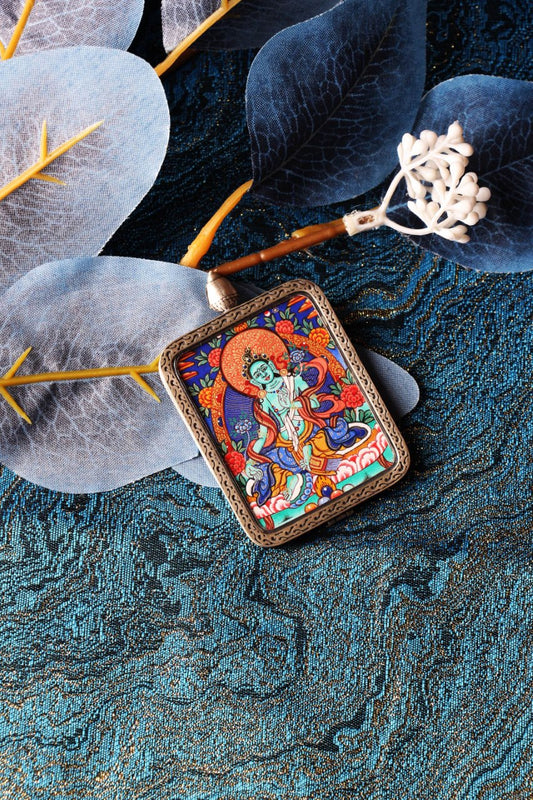 Angled flat lay of a Green Tara mini thangka pendant in a silver-plated case on a blue textile background