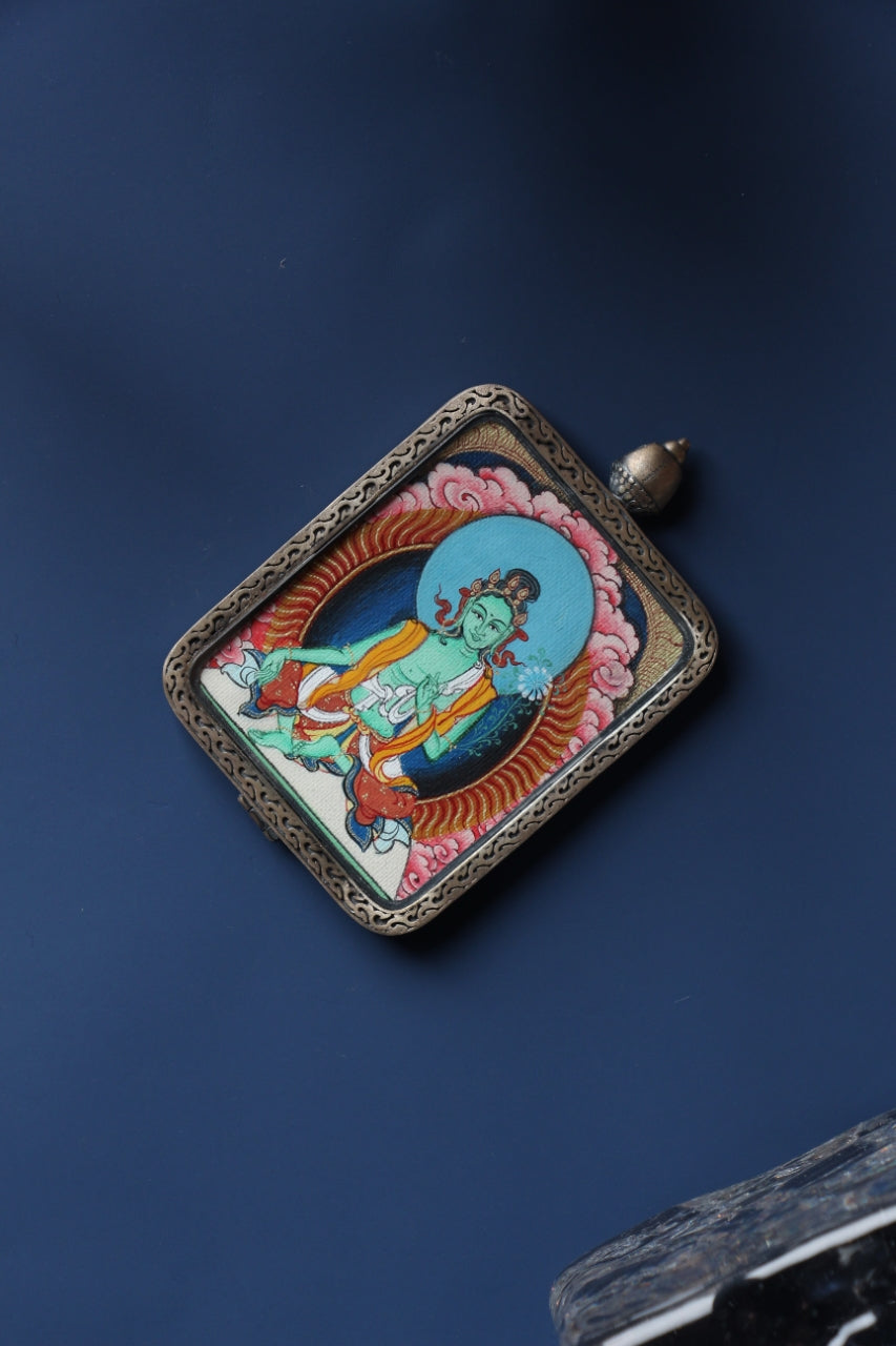 Angled product view of the Green Tara mini thangka pendant from the Tibetan Red Cloud Zangka Series, fully hand-painted in Tibet with natural mineral pigments and presented in a Dunhuang Nine-Colored Deer sterling silver case for portable devotion and collecting.