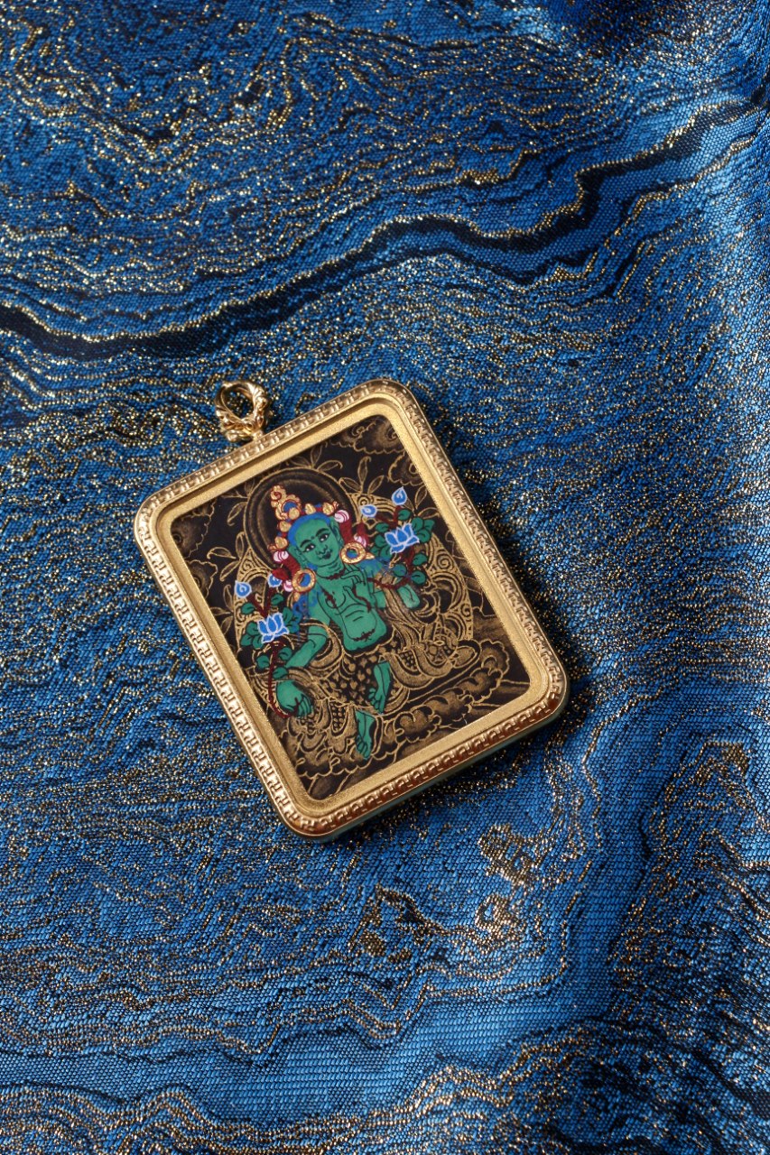 Green Tara black-gold miniature thangka pendant in a gold-tone titanium steel case, photographed on traditional blue brocade fabric