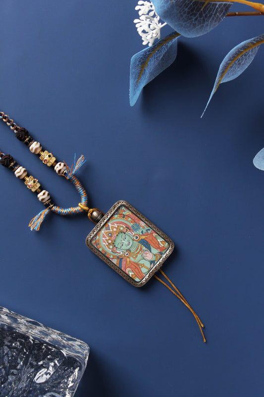 Flat lay of Green Tara mini thangka pendant with braided necklace, sterling silver case, and tassel cords on a dark blue background