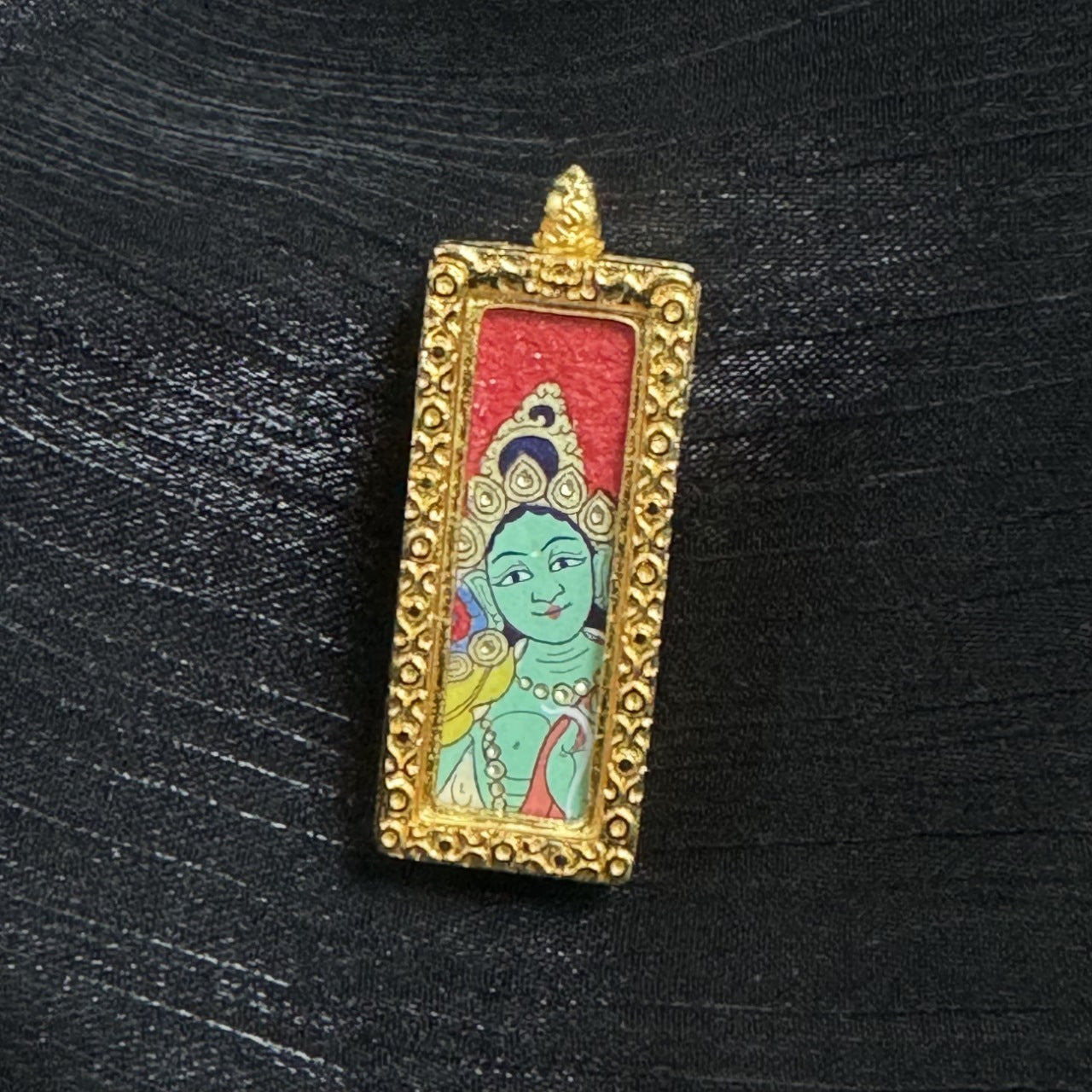 Front close-up of a Green Tara mini thangka pendant showing the green face, crown motif, necklace and garment color accents, red background, and the gold-tone raised frame around the painting.