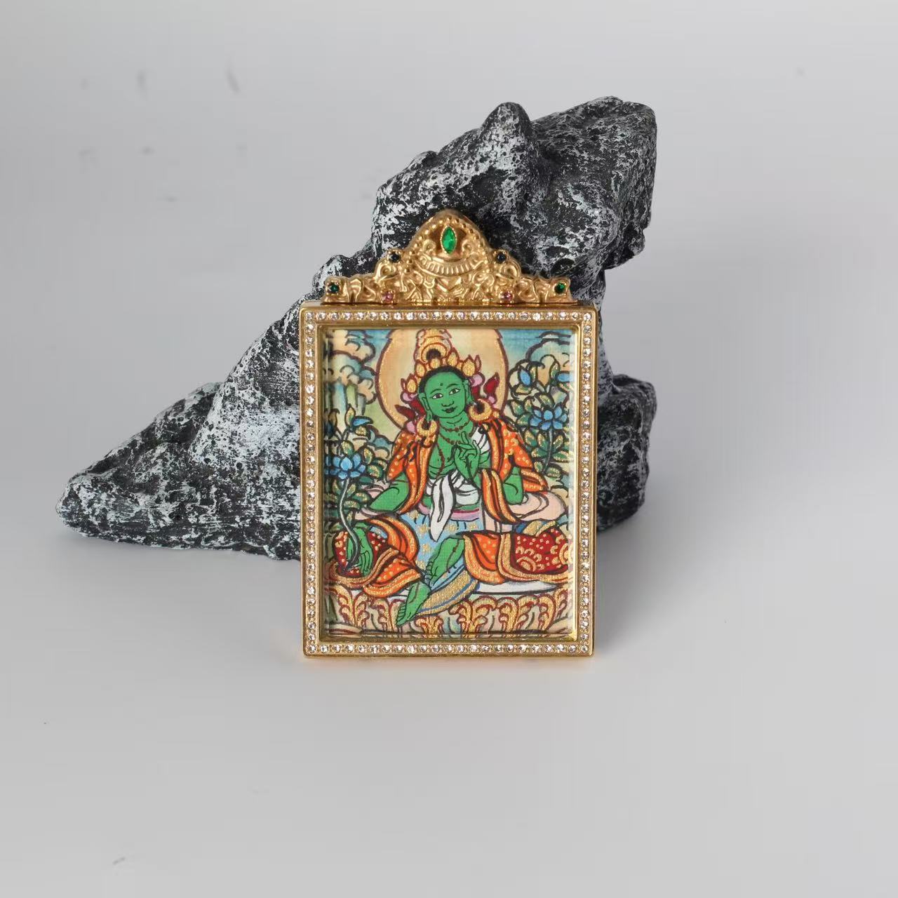 Front view of Green Tara mini thangka pendant showing the hand-painted Green Tara figure, gold-tone rectangular frame with crystal border, and ornate top crown with a green gemstone, photographed on a rock prop.