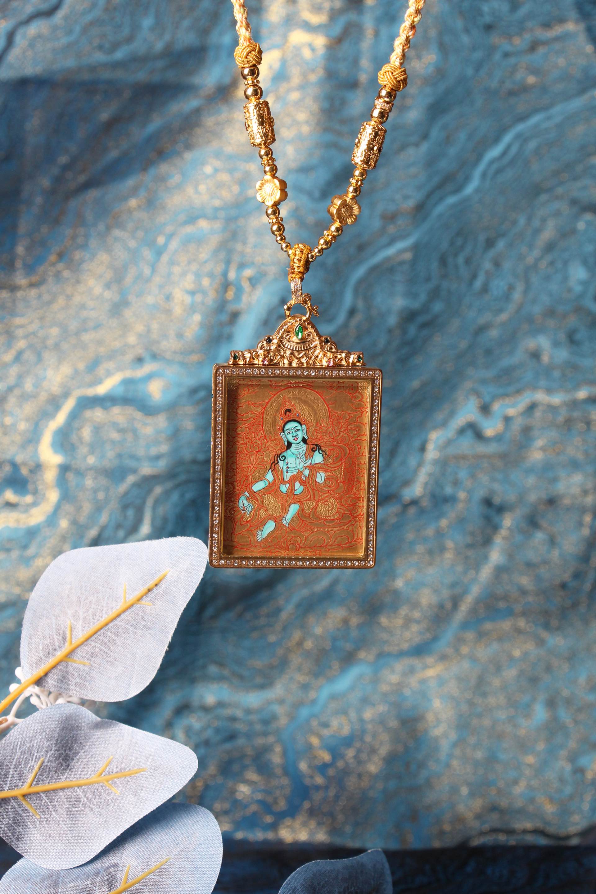 Green Tara gold-ground mini thangka pendant shown hanging on beaded necklace, square crystal-bordered frame, ornate top setting with green gemstone, decorative background display