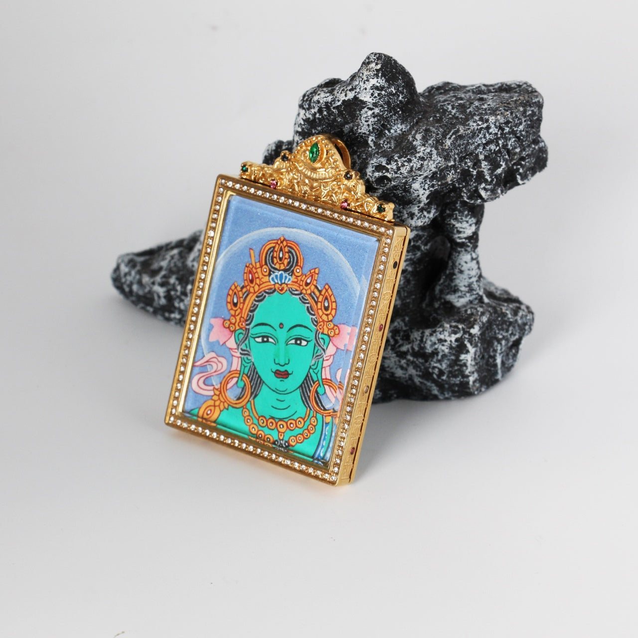 Angled view of Green Tara Buddha head mini thangka pendant showing the raised titanium steel housing, crystal-style bezel border, and decorative crown-like top with colored accents