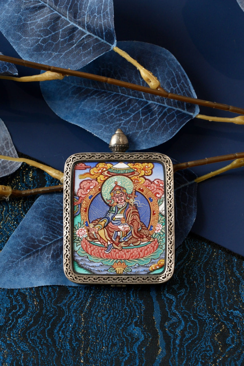 Hand-painted Guru Rinpoche seated mini thangka pendant set in a silver gawu case displayed against blue leaves and textile background.