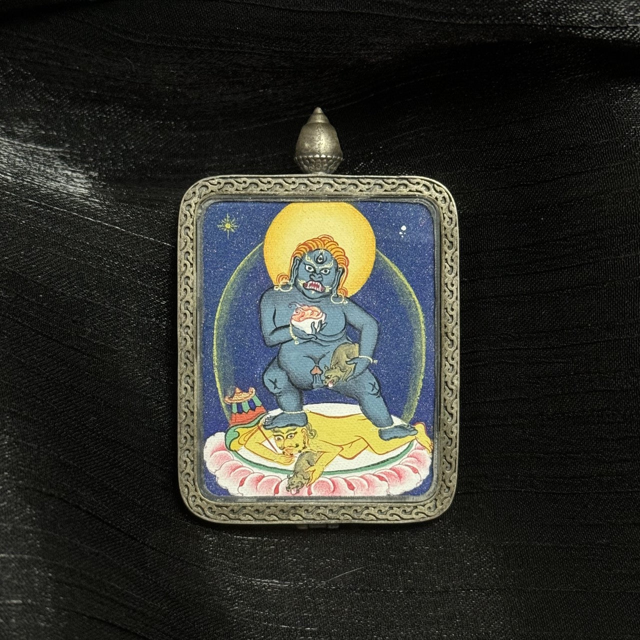 Close-up of Black Jambhala mini thangka pendant showing hand-painted deity details, mineral pigment layering, and traditional silver casing