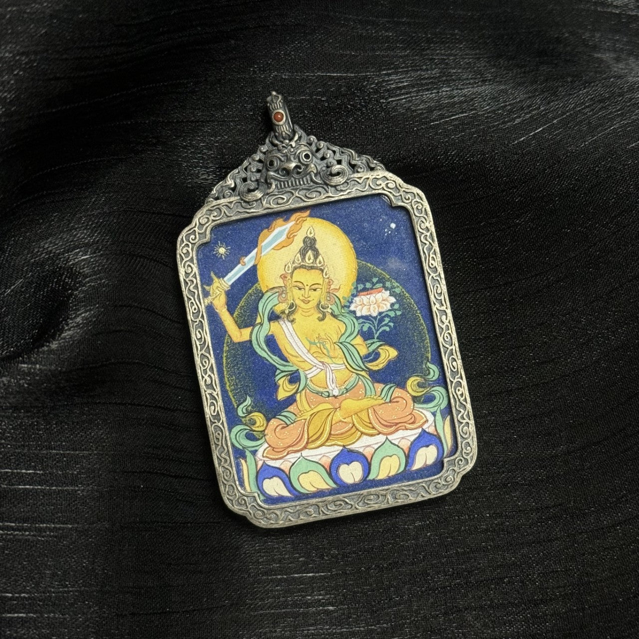 Angled view of Manjushri mini thangka pendant highlighting mineral pigment layering, flowing silk ribbons, lotus pedestal, and antique silver frame craftsmanship.