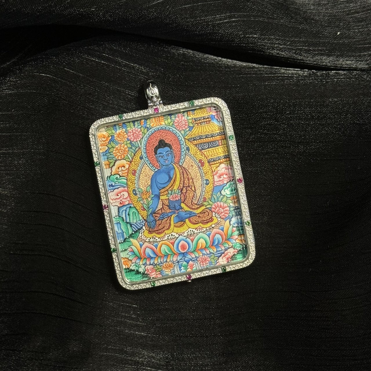 Close-up view of a hand-painted Medicine Buddha mini thangka pendant featuring vibrant mineral pigments, traditional lotus throne, halo, and ornate floral background details.