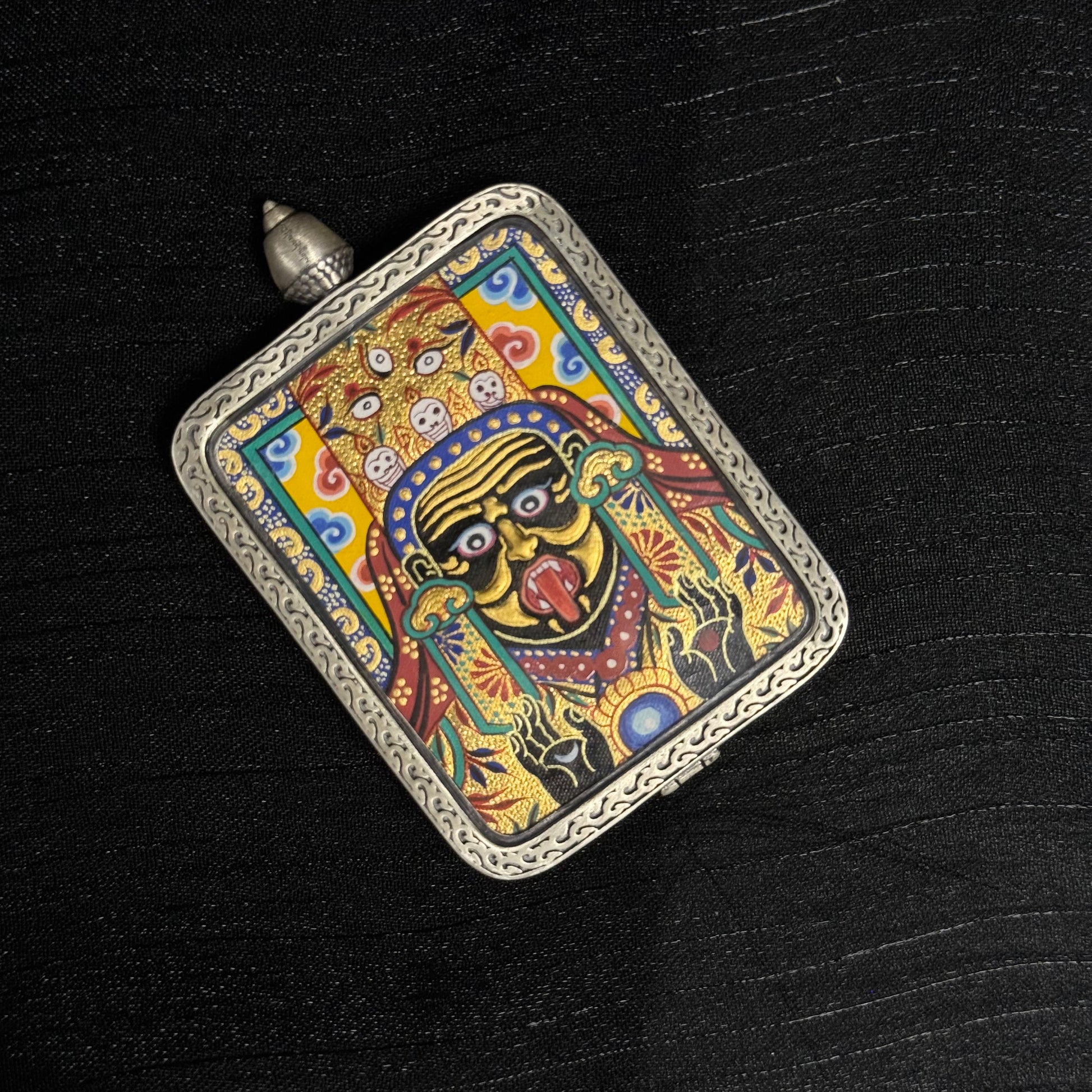 Top view of hand-painted Zaki Lhamo miniature thangka pendant displaying layered gold powder highlights, colorful traditional thangka composition, and silver protective frame.