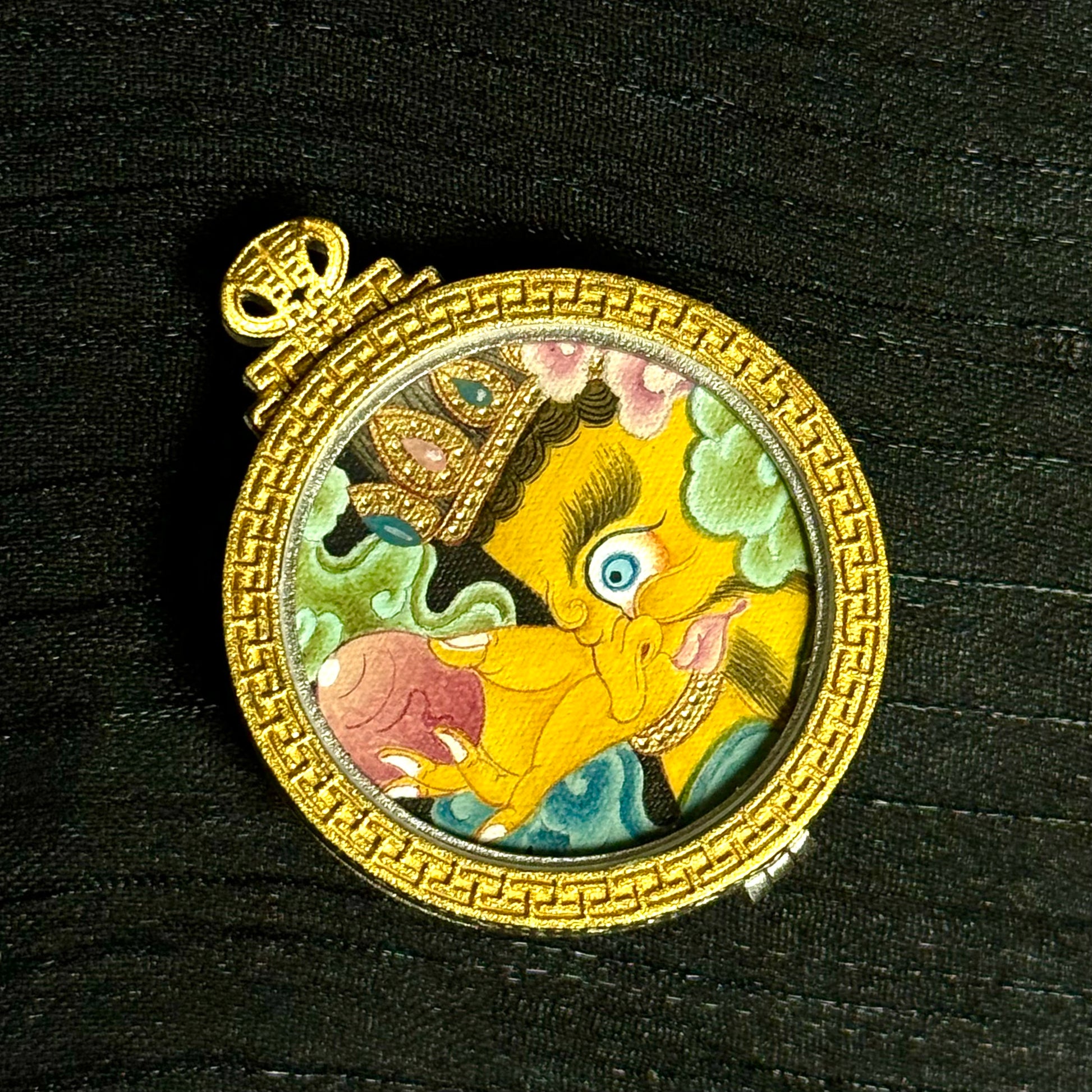 Close-up view of the circular Yellow Jambhala thangka painting inside the pendant, highlighting the deity’s side-profile face, crown ornament, wish-fulfilling jewel, and layered mineral pigment colors.