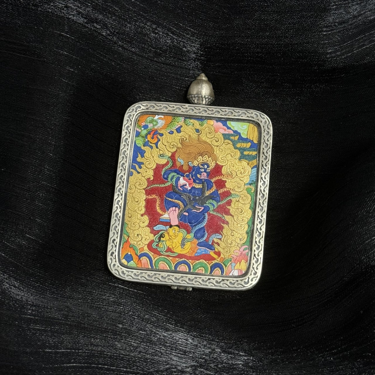 Close-up front view of a Black Jambhala mini thangka amulet pendant in a carved silver frame, depicting the wealth deity stepping on a figure, surrounded by vibrant mineral pigment colors and ornate golden aura.
