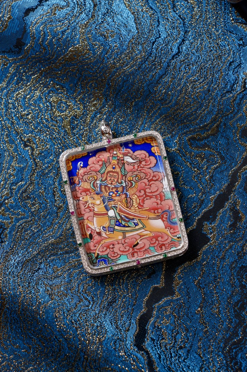 Front view of King Gesar hand-painted mini thangka pendant in a 925 silver case, photographed on blue brocade fabric.