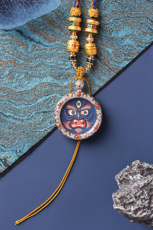 Front view of a Mahakala mini thangka pendant featuring a fierce protector face, hand-painted with rough mineral pigments, set in a jewelry-grade copper case with decorative beads and braided cord.