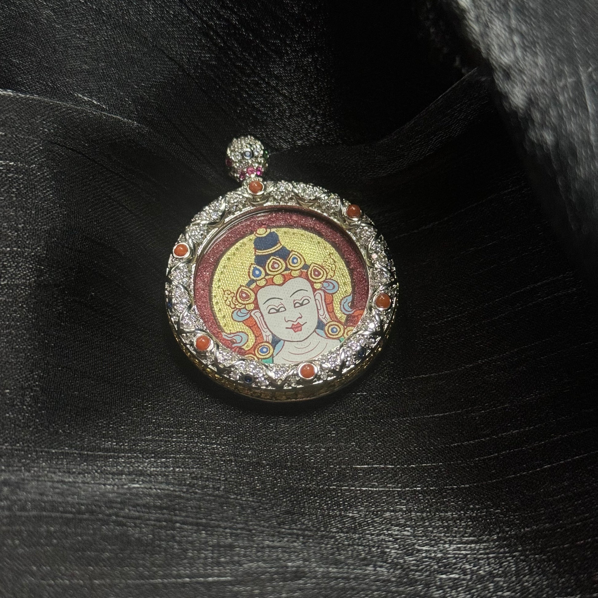 Mahasthamaprapta Bodhisattva miniature thangka pendant resting on dark silk fabric highlighting hand-painted Buddha head, gold halo, and gemstone-set copper frame