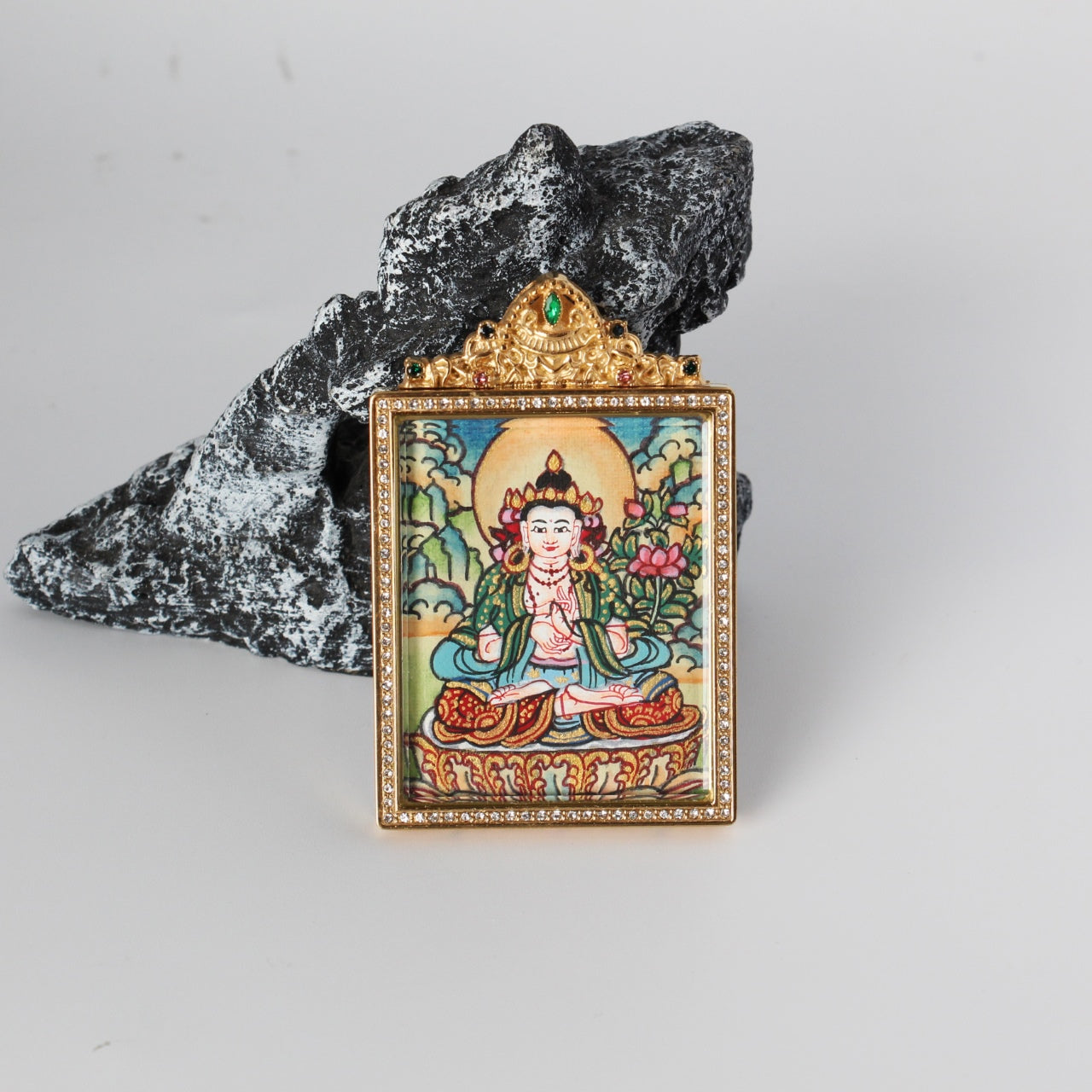 Front view of Mahasthamaprapta Bodhisattva mini thangka pendant showing the hand-painted seated bodhisattva with halo and lotus motif, gold-tone rectangular frame with crystal border, and ornate crown top with a green gemstone, photographed on a rock prop.