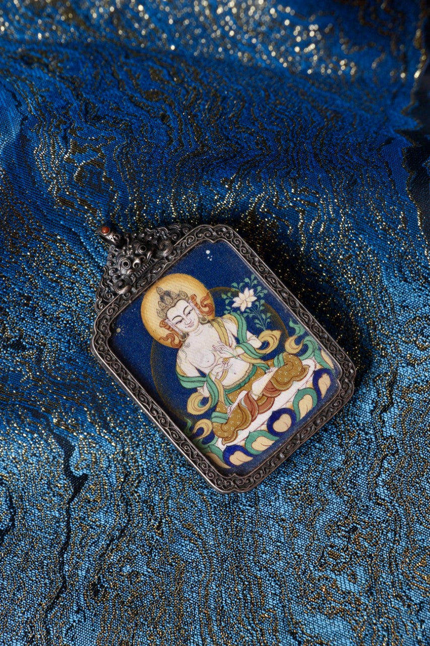 Mahasthamaprapta Bodhisattva mini thangka pendant displayed on blue brocade, featuring a hand-painted image with mineral pigments in a vintage silver setting.