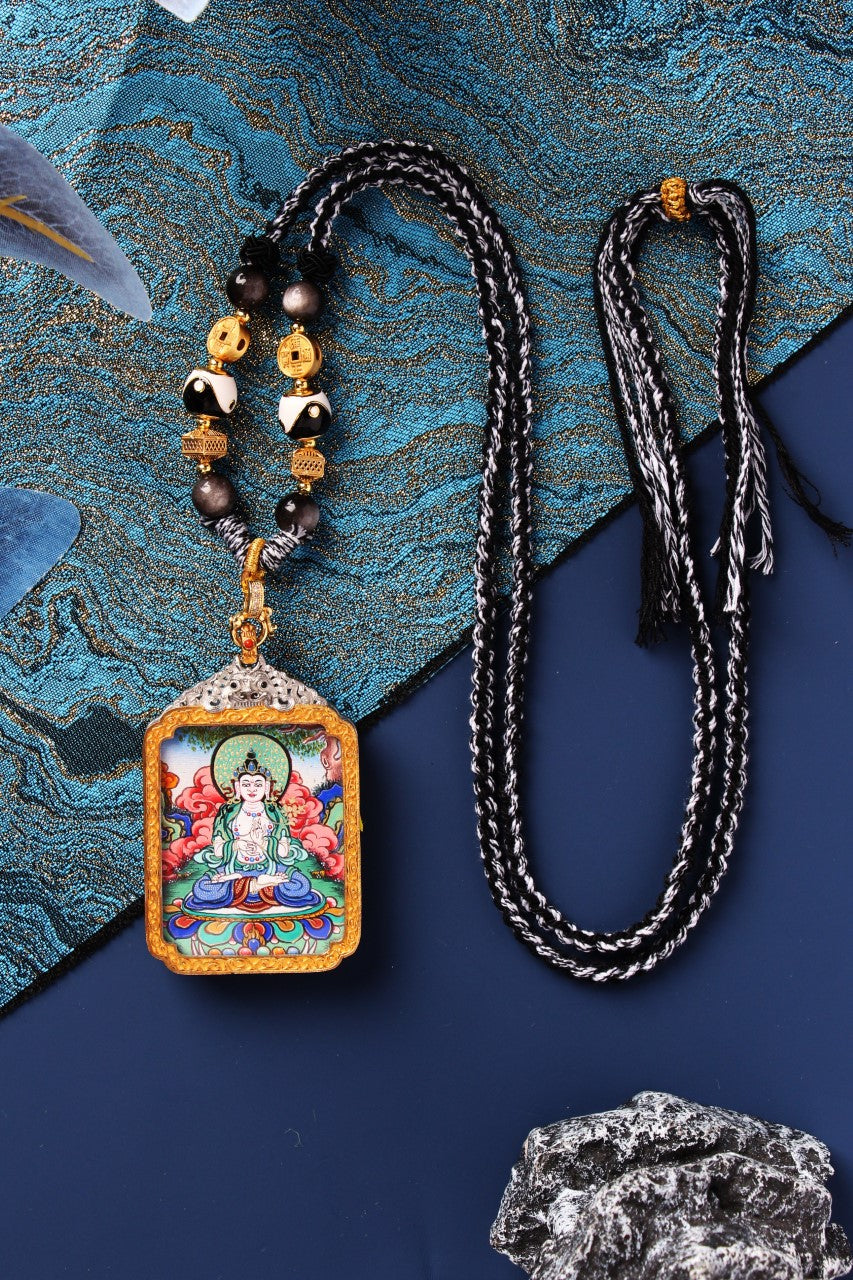 Flat lay of Mahasthamaprapta Bodhisattva mini thangka pendant in a silver-plated case with gold-tone frame, shown with beaded necklace styling and a long black-and-white braided cord, placed on textured blue brocade fabric with rock accent.