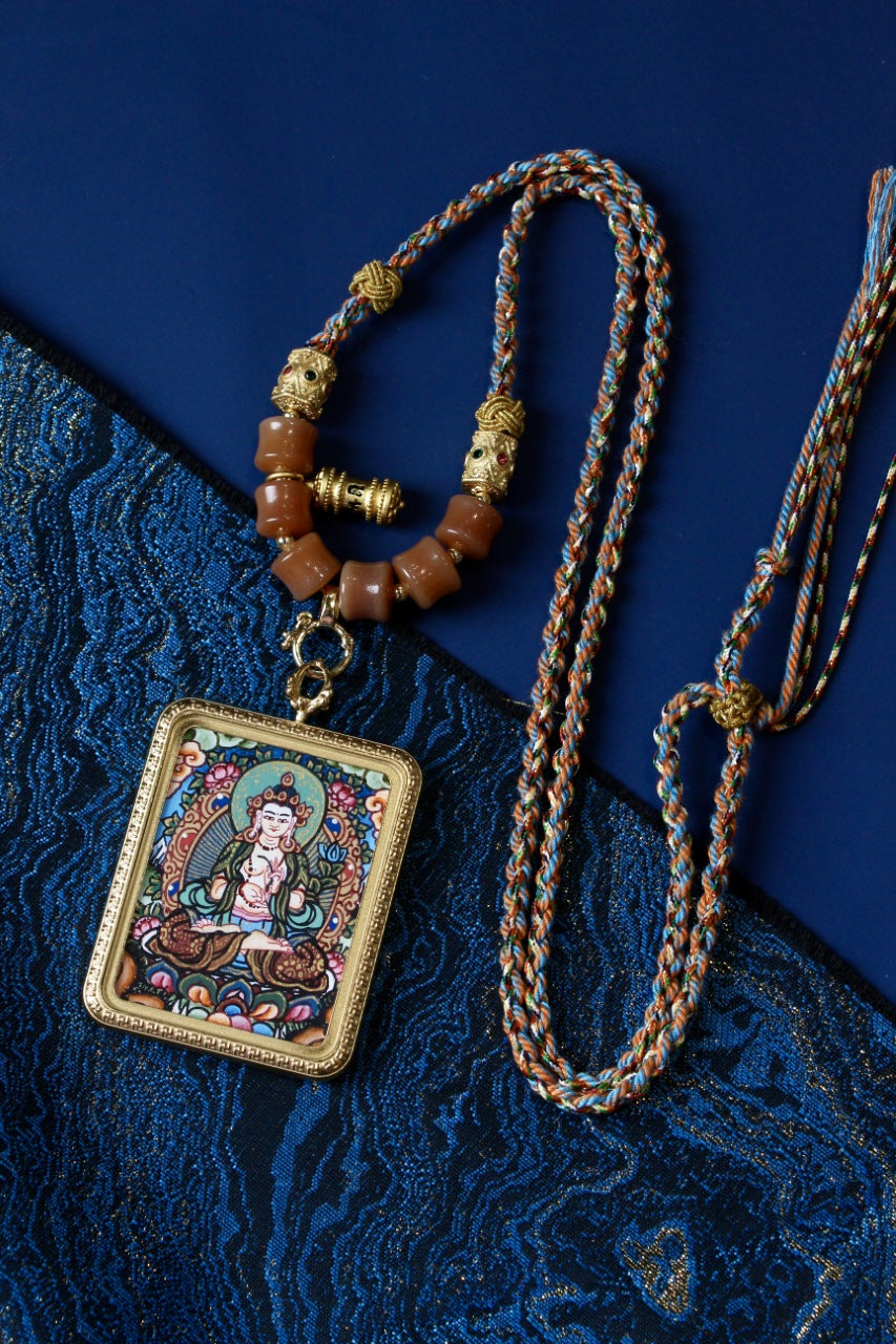 Flat lay of a Mahasthamaprapta Bodhisattva Nepal thangka pendant necklace with multicolor braided cord, beads, and gold-tone accents on a blue background.