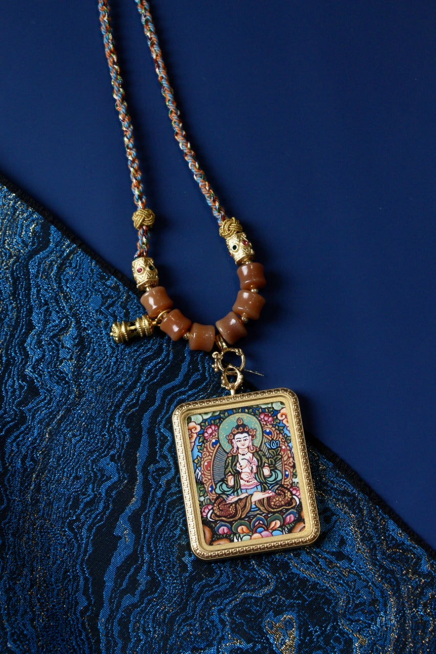 Mahasthamaprapta Bodhisattva hand-painted Nepal thangka pendant necklace shown hanging with braided cord and bead details against a blue textile background.