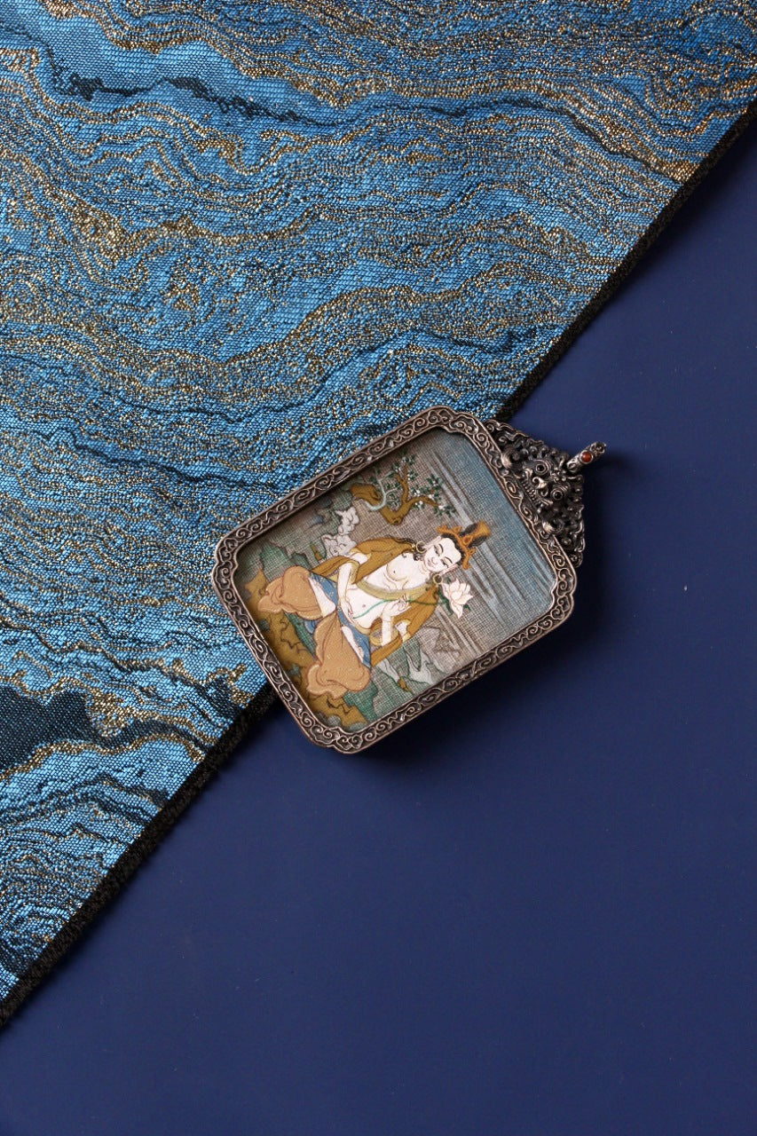 Angled view of Mahasthamaprapta Bodhisattva hand-painted mini thangka pendant in vintage silver gawu case on blue textile background
