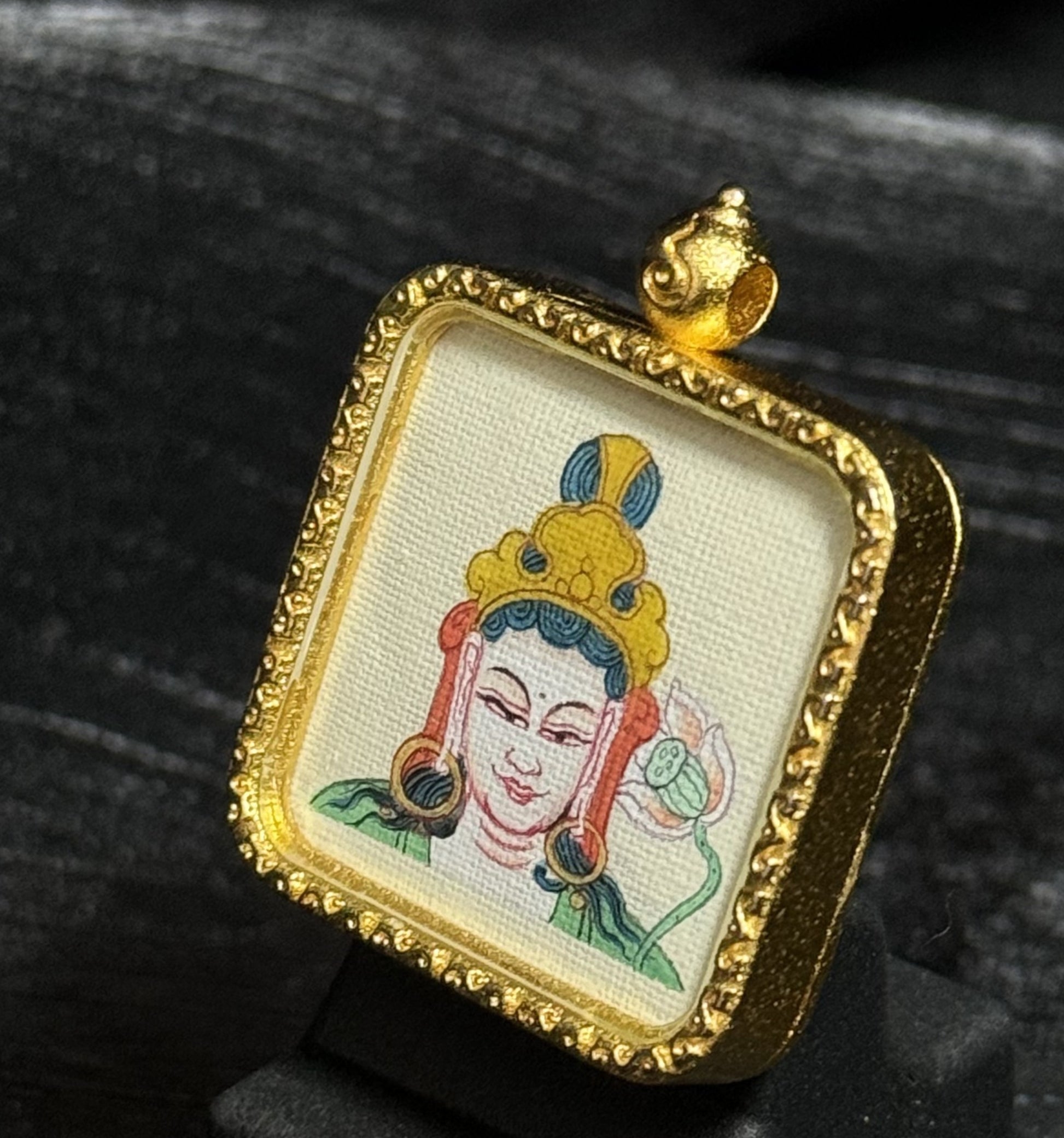 Mahasthamaprapta Bodhisattva mini thangka pendant displayed on dark fabric background, showing hand-painted deity portrait with crown and lotus painted on canvas inside a decorative gold-toned pendant frame.