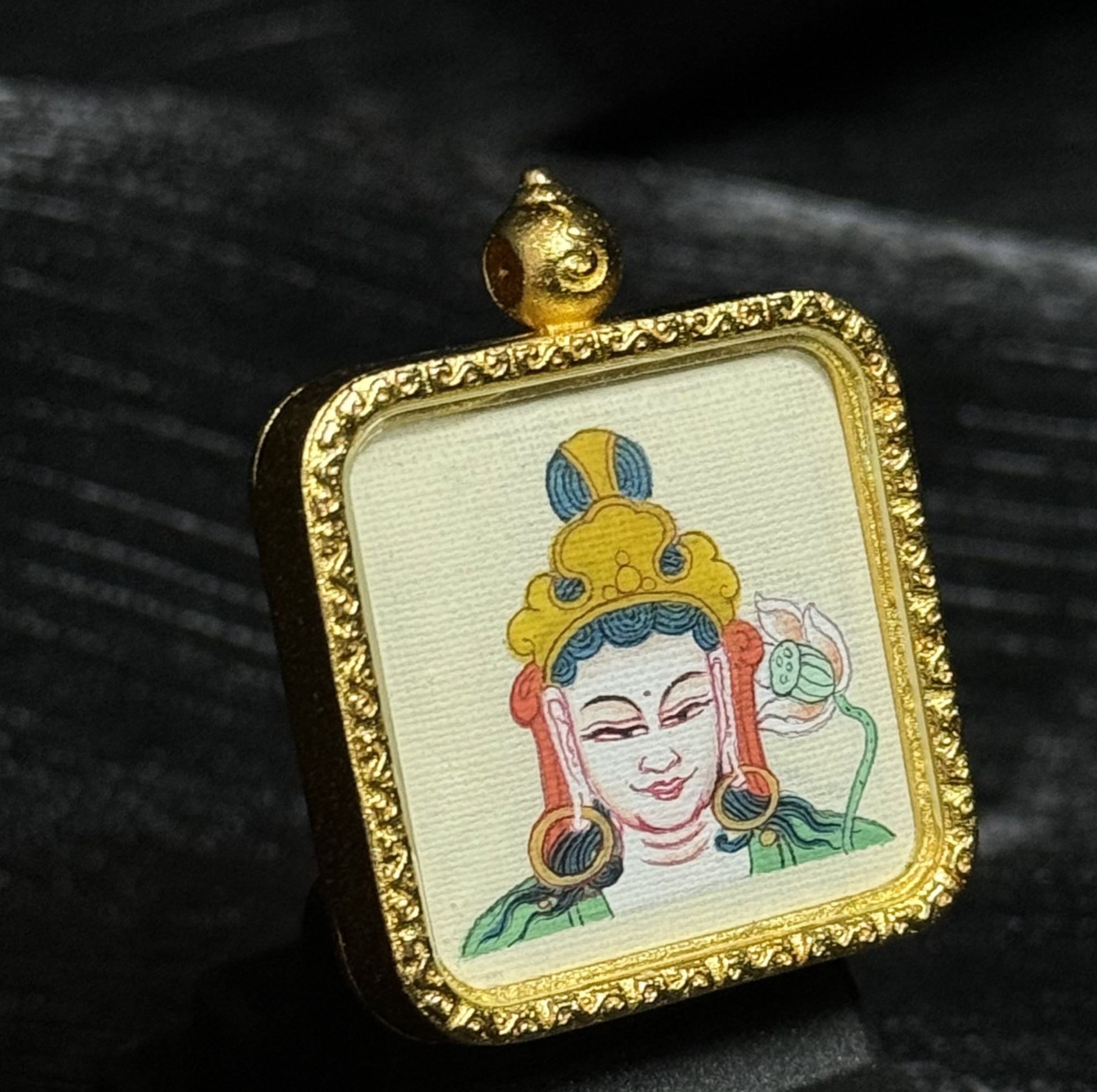 Angled view of a Mahasthamaprapta Bodhisattva miniature thangka pendant showing detailed hand-painted deity portrait on textured canvas within a gold-toned framed pendant setting.
