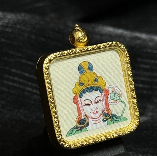 Angled view of a Mahasthamaprapta Bodhisattva miniature thangka pendant showing detailed hand-painted deity portrait on textured canvas within a gold-toned framed pendant setting.