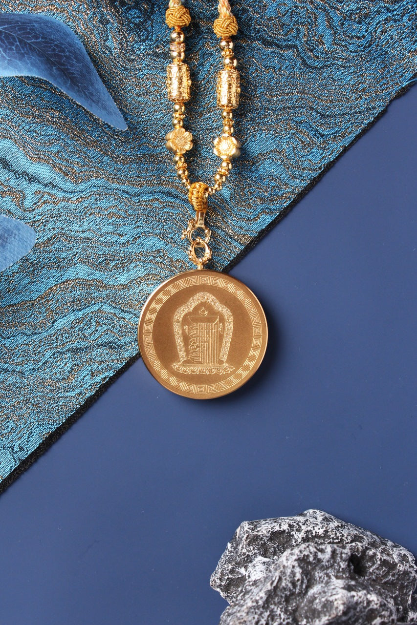 Back view of a round titanium steel pendant case engraved with a sacred symbol and geometric border, connected to a braided necklace with gold-tone bead elements.