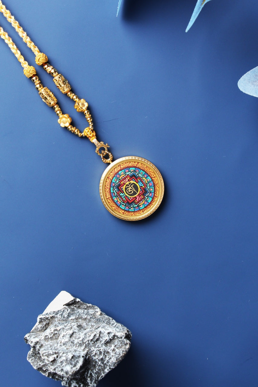 Diagonal flat-lay photo of a mandala mini thangka pendant featuring vivid mineral pigment colors, round gold-tone titanium steel case, braided cord, and decorative beads on a dark blue surface.
