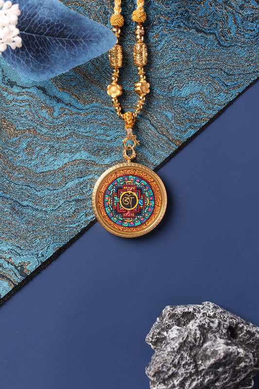 Front view of a round mandala mini thangka pendant, hand-painted with multicolor geometric patterns and Sanskrit seed syllable at the center, set in a gold-tone titanium steel case with decorative bead necklace.