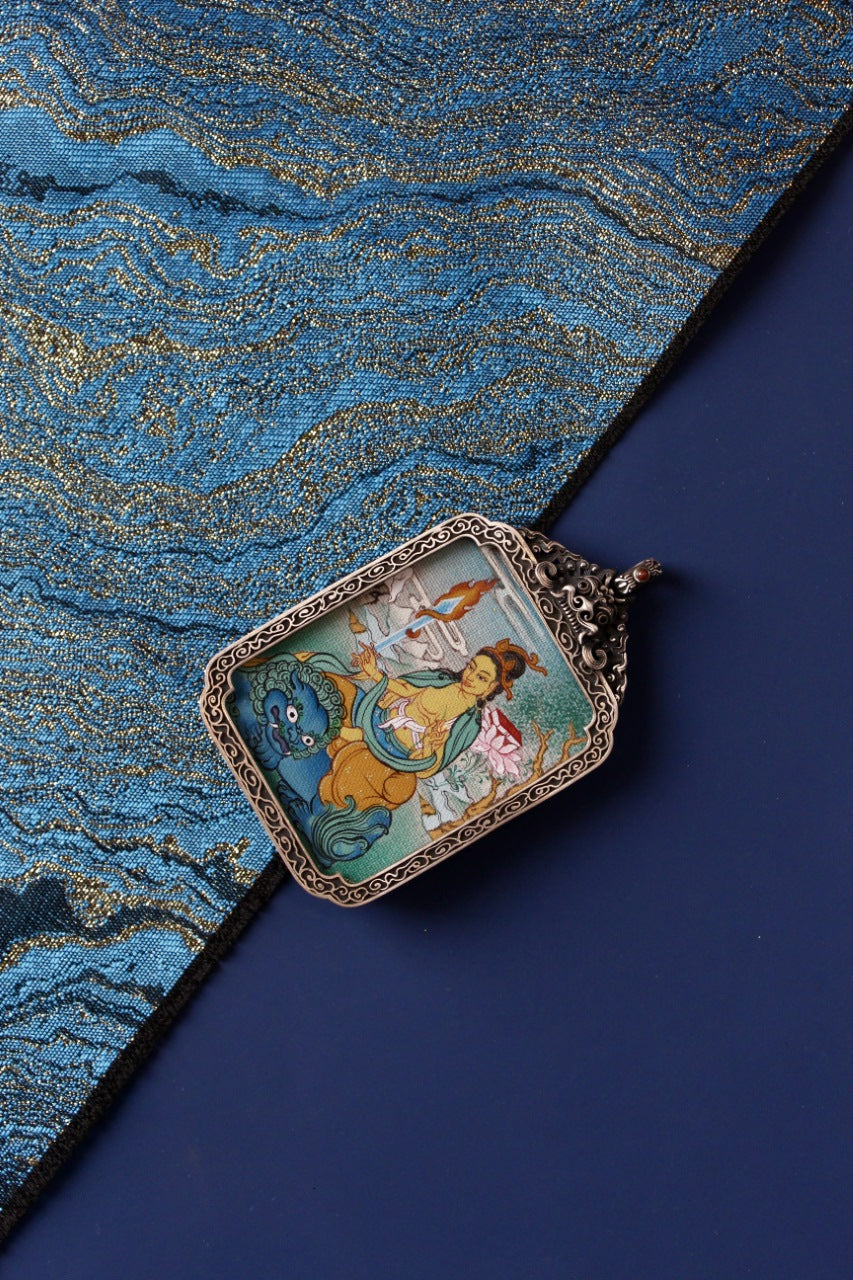 Angled flat lay of Manjushri Bodhisattva mini thangka pendant on traditional textile background, showing the hand-painted thangka artwork in a vintage silver case