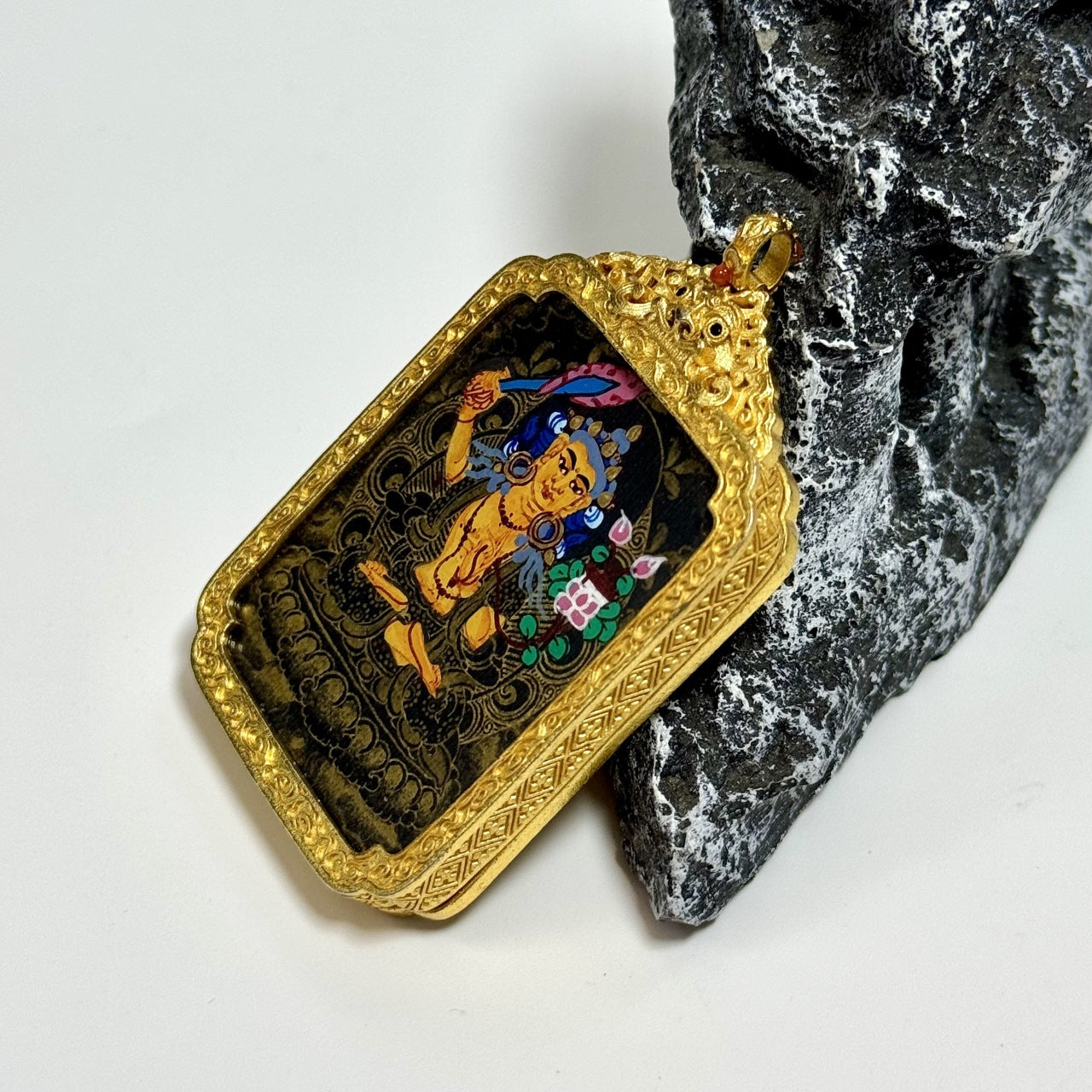 Angled view of Manjushri Bodhisattva mini thangka pendant, hand-painted with mineral pigments in Nepal black gold style, mounted in an ornate copper protective case