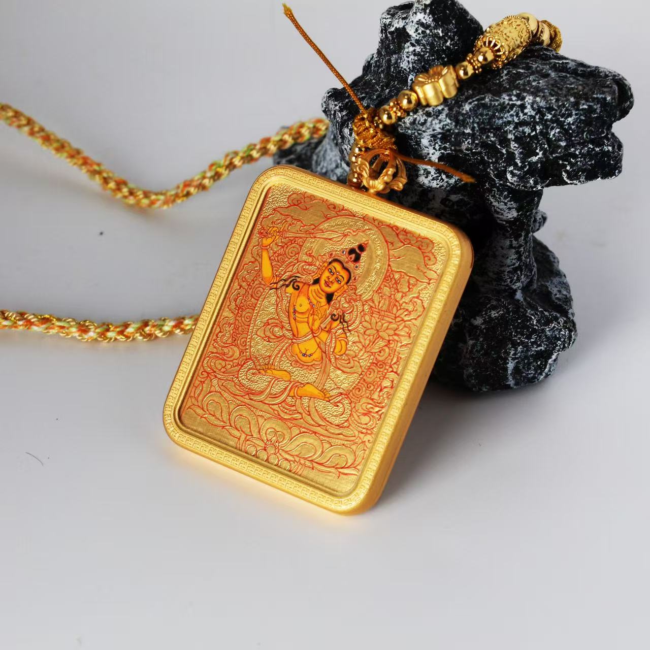 Close view of a Manjushri Bodhisattva mini thangka pendant showing the braided cord, decorative metal beads, and hanging connector above the copper-framed gold-ground painting.