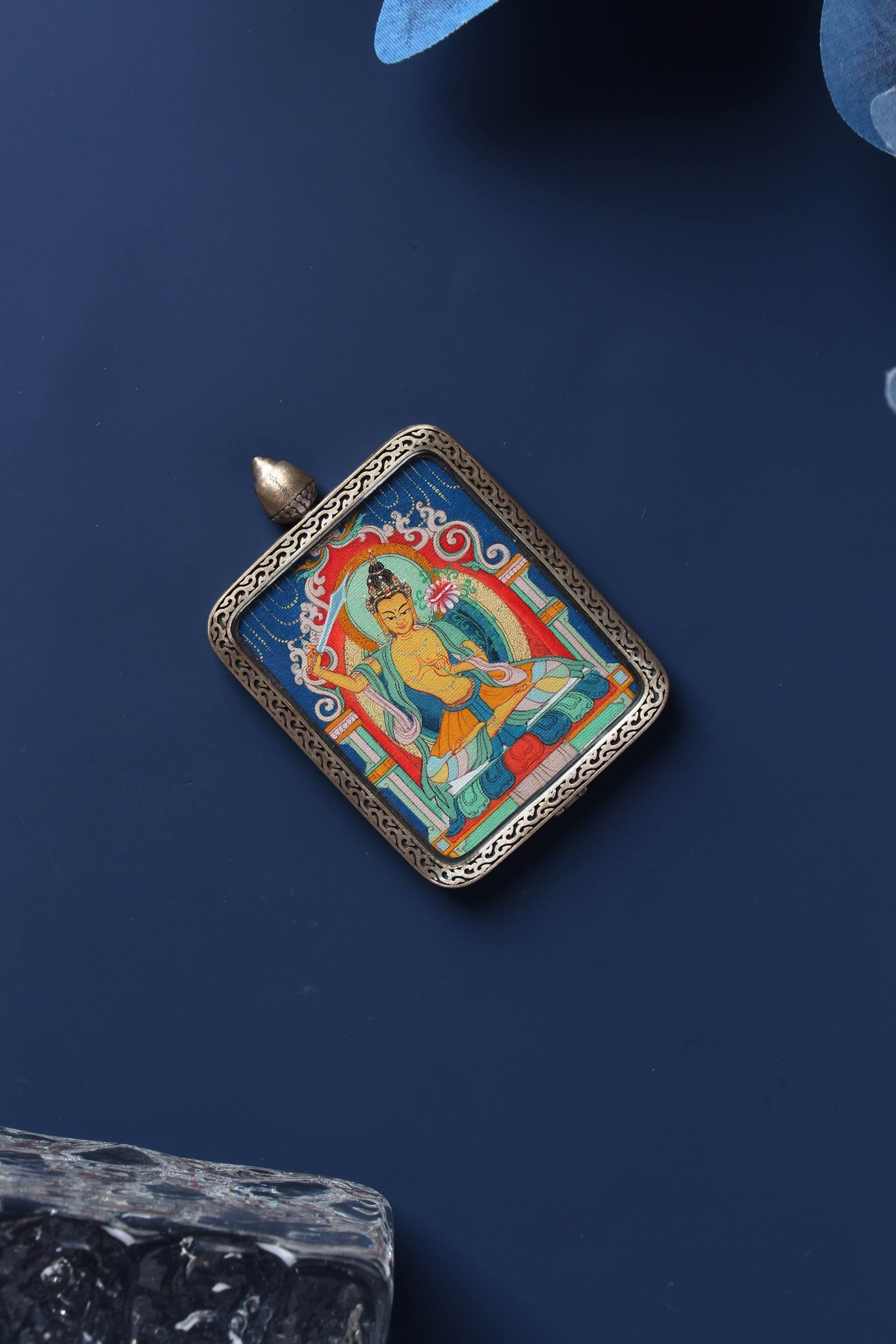 Flat lay of a hand-painted Manjushri Bodhisattva mini thangka pendant in a Dunhuang Nine-Color Deer sterling silver case on a dark blue background