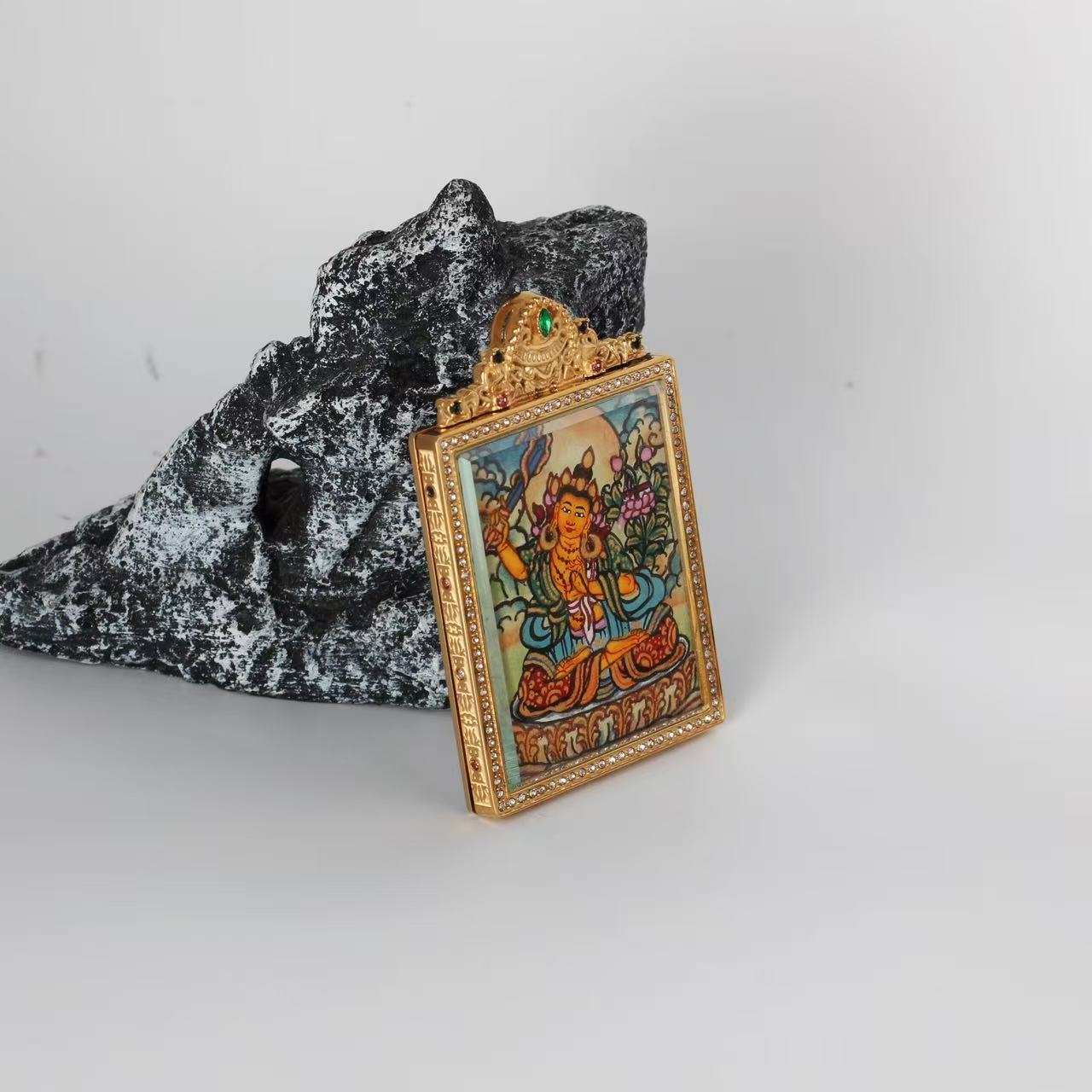 Side-angle view of the Manjushri Bodhisattva mini Thangka pendant showing the ornate crown-shaped top with colored stones, the crystal border, and the gold-tone titanium steel frame.