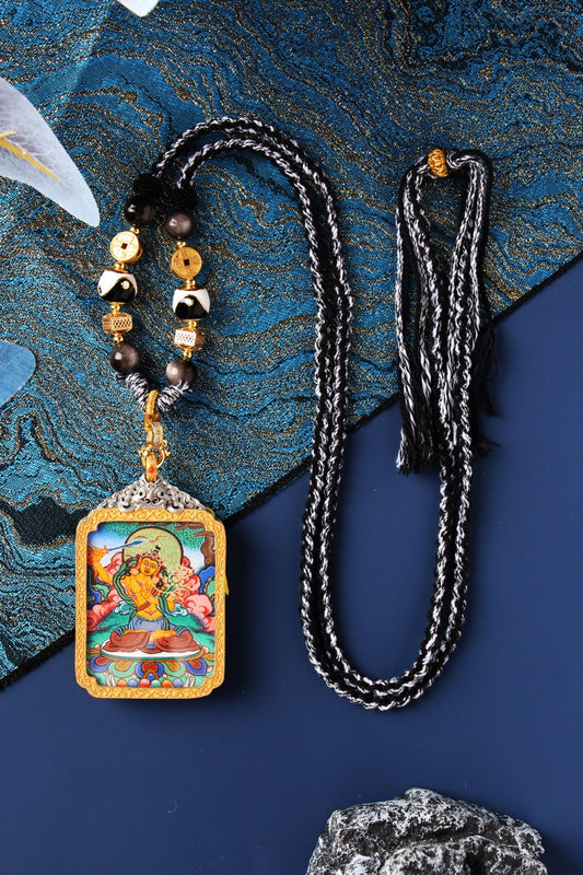 Flat lay of Manjushri Bodhisattva mini thangka pendant in a silver-plated case with gold-tone frame, shown attached to a decorative beaded necklace with coin-like gold accents and black-and-white beads, alongside a long black-and-white braided cord, arranged on textured blue brocade fabric with a stone prop.