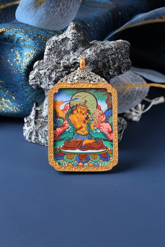 Front view of Manjushri Bodhisattva mini thangka pendant, fully hand-painted with mineral pigments, showing the deity with raised sword gesture, seated on a lotus base with multicolor detailing, framed by a gold-tone border and ornate silver-plated top plate, photographed with dark stone and blue brocade fabric in the background.