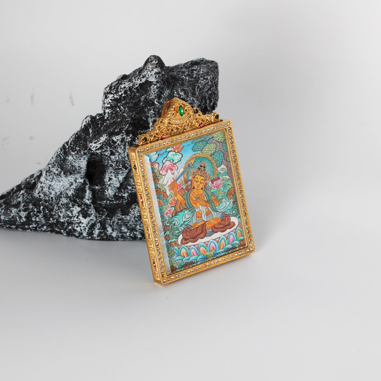 Close angled view of a hand-painted Manjushri Bodhisattva thangka pendant, featuring vivid turquoise and gold tones inside a crystal-studded frame.