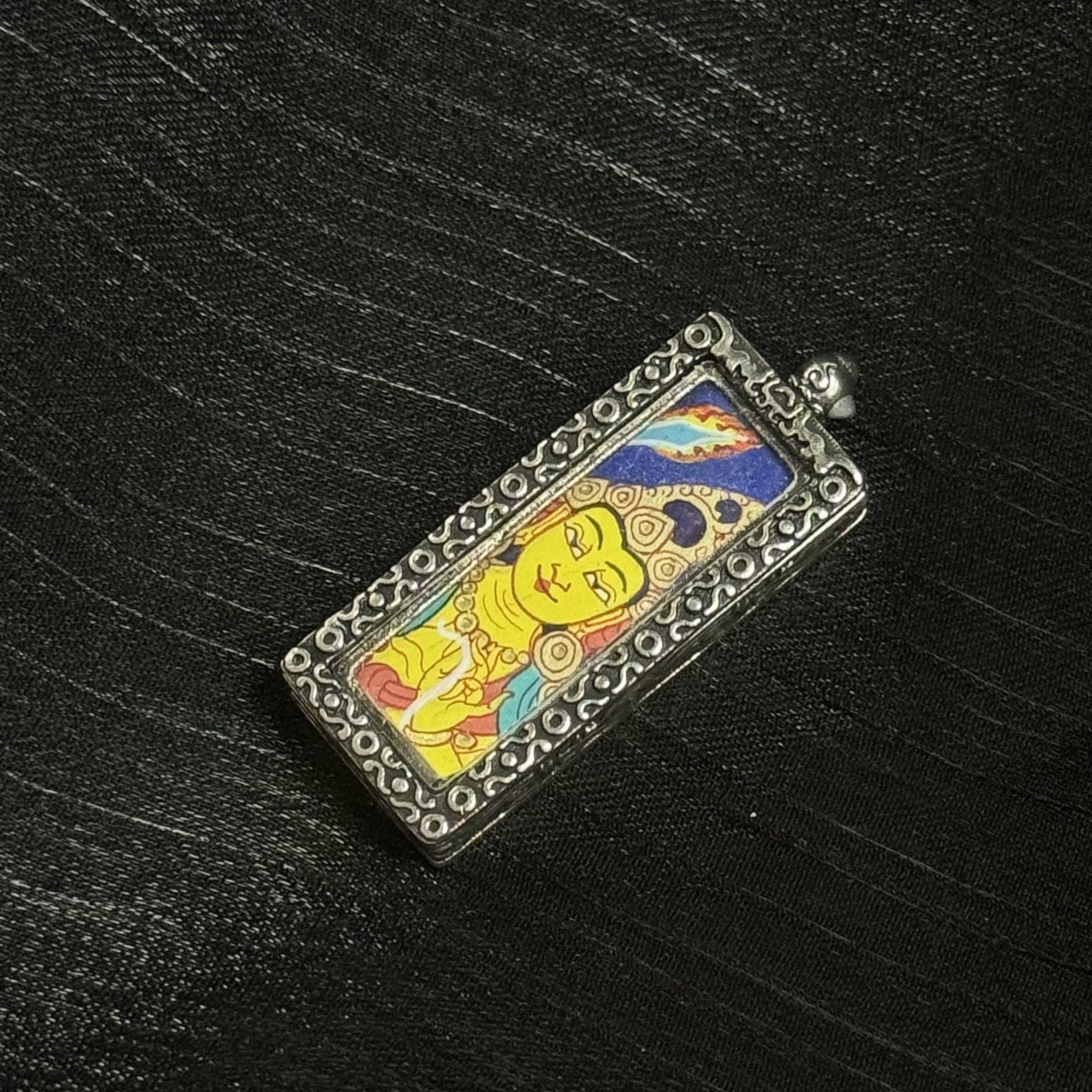 Angled view of a Manjushri mini thangka pendant highlighting the slim rectangular pendant shape, raised decorative metal border, and the vivid blue-and-gold color contrast inside the frame.