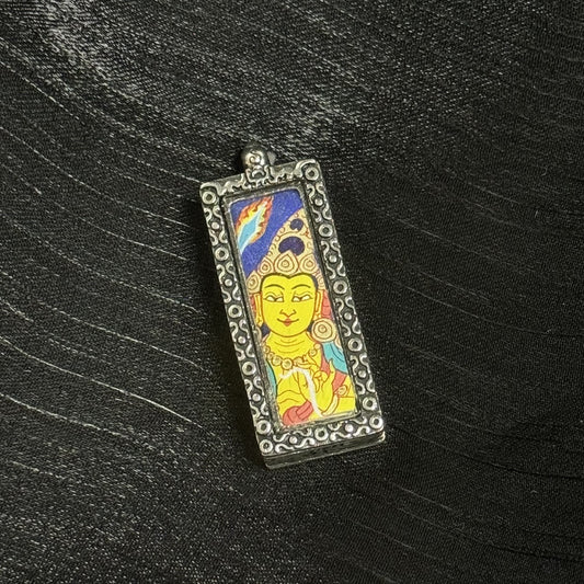 lose-up of the Manjushri mini thangka pendant showing fine linework on the eyes and lips, layered mineral-pigment color blocks, and the textured ornamental metal frame around the painting.