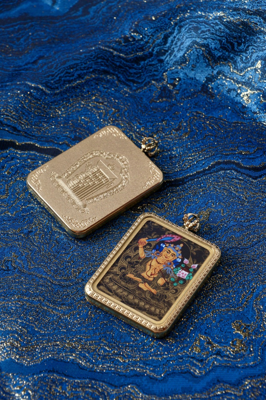 Flat-lay image of a Manjushri Mini Thangka Pendant displayed together with its titanium steel case. The artwork features the distinctive deep black background and hand-applied gold detailing characteristic of Nepalese black-gold thangka art. The pendant measures 3.5 × 4.5 cm and is photographed on a deep blue brocade textile, emphasizing traditional craftsmanship and material quality.