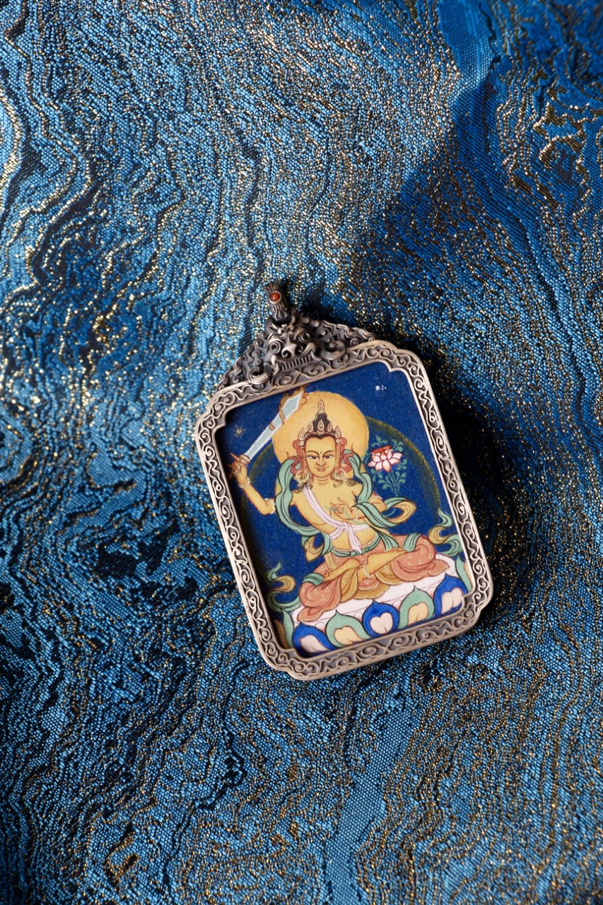 Flat-lay close-up of a Manjushri Bodhisattva thangka pendant on blue brocade, featuring mineral-pigment painting and a detailed silver frame.