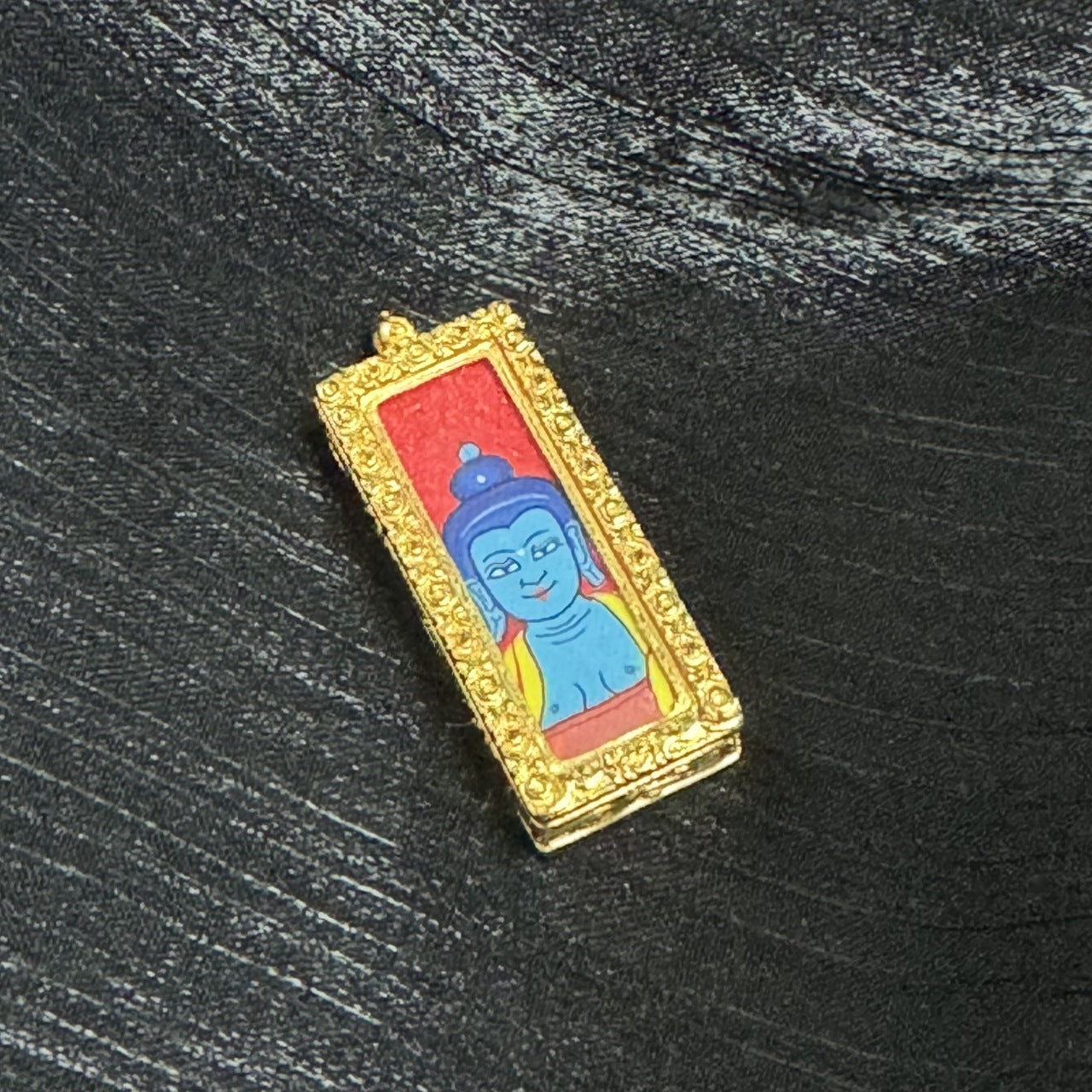Angled view of a Medicine Buddha mini thangka pendant highlighting the slim rectangular form, the textured gold-tone ornamental frame, and the red-ground Buddha head painting inside the copper case.